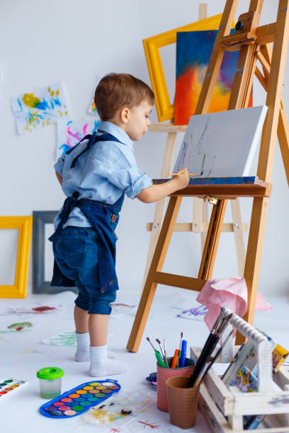 かわいい、深刻で集中した、青いシャツとジーンズのエプロンを着た白い3歳の男の子が、イーゼルの上に立ってキャンバスに描いています。幼児教育、才能、幸せな家族や子育ての概念 - artists canvas indoors childhood small ストックフォトと画像