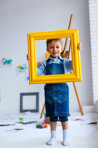 Cute, happy, white boy in blue shirt and jeans smiling and looking through yellow frame. Little child having fun in artist studio. Concept of early childhood education, happy family, parenting