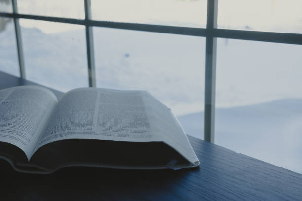 Open bible on a dark wood table by a window with grill inserts with a landscape covered in snow outside