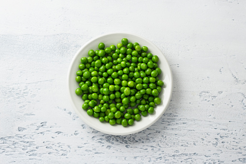 White plate with steamed green peas on a light blue background, flat lay. Healthy delicious dietary ingredient for cooking