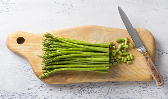 Wooden board of fresh asparagus with cut tough pieces and a knife on a light blue background, home cooking, top view