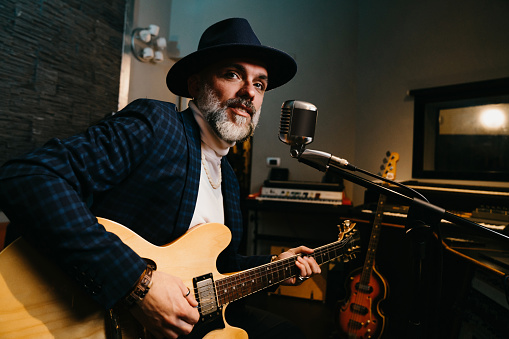 A musician is playing guitar and singing in a professional recording studio. He's wearing a cool hat while he's playing his electric guitar.