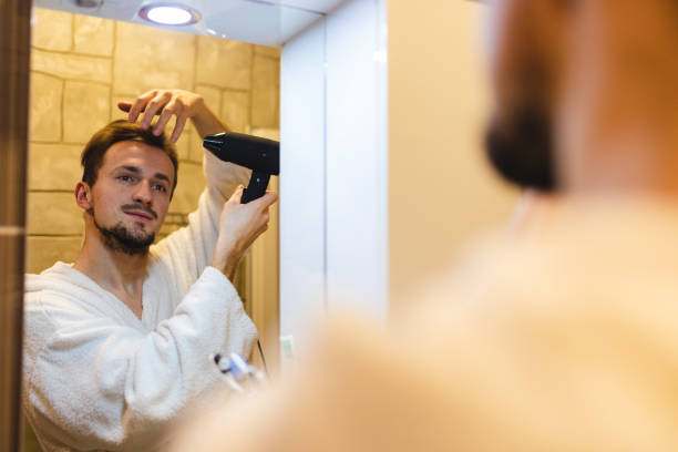 jeune homme souriant en peignoir se séchant les cheveux après avoir pris une douche tout en se regardant dans le miroir de la salle de bain - toothbrush brushing teeth brushing dental hygiene photos et images de collection