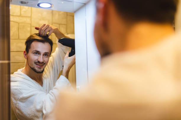 jeune homme souriant en peignoir se séchant les cheveux après avoir pris une douche tout en se regardant dans le miroir de la salle de bain - brushing teeth photos et images de collection