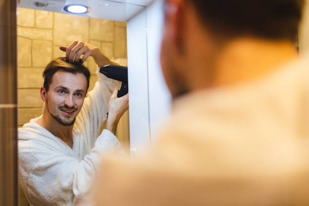 istock Smiling young man in a bathrobe blow drying his hair after taking a shower while looking at himself in the bathroom mirror 1362982645