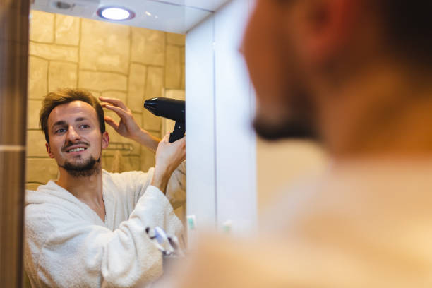 istock Smiling young man in a bathrobe blow drying his hair after taking a shower while looking at himself in the bathroom mirror 1362982646