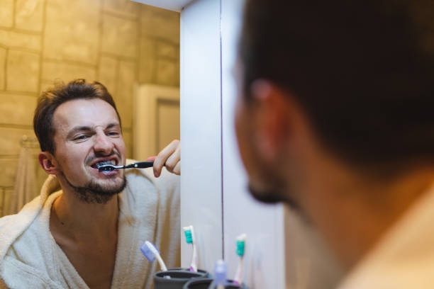 jeune homme en peignoir se brossant les dents tout en se regardant dans le miroir de la salle de bain - toothbrush brushing teeth brushing dental hygiene photos et images de collection