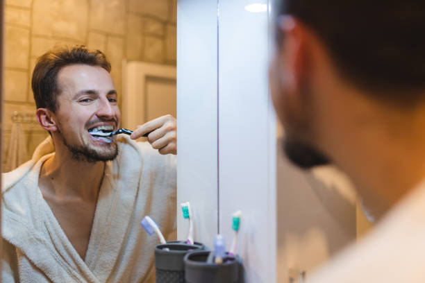 jeune homme en peignoir se brossant les dents tout en se regardant dans le miroir de la salle de bain - toothbrush brushing teeth brushing dental hygiene photos et images de collection