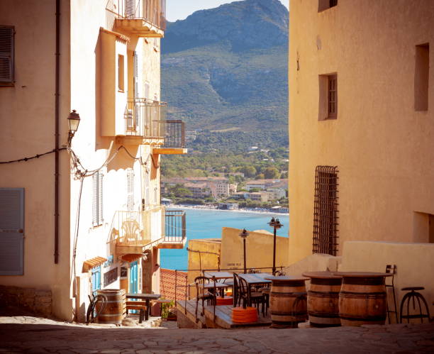 Calvi, Corsica, France - September, 2019: Mountains, beach, boats and port of Calvi as shown in background through a tunnel view of the historic old city's citadel narrow streets where restaurants are closed for the siesta