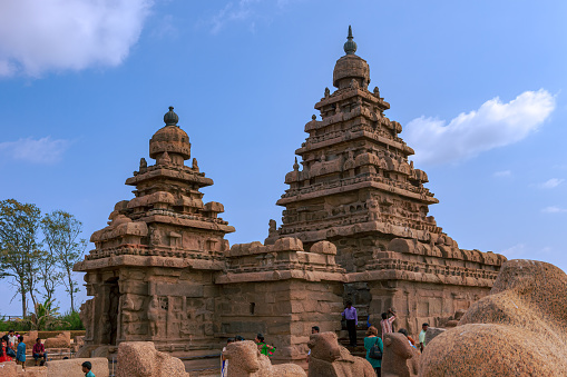 Mahabalipuram, India - January 30, 2015: Some tourists both local Indian and foreigners at the one thousnd three hundred yar old Shore Temple in Mahabalipuram or Mamallapuram, built in the 8th Century, between 700 and 728 AD, during the rign of the Pallava King Narasimhavarman II.  Declared a UNESCO World Heritage site in 1984, the temple is one of the oldest structural temples in South India. It survived the Tsunami of 2004. The sides display sand and wind errosion over 1300 years. Photo shot around noon against a blue sky with some clouds.  No people; copy space.
