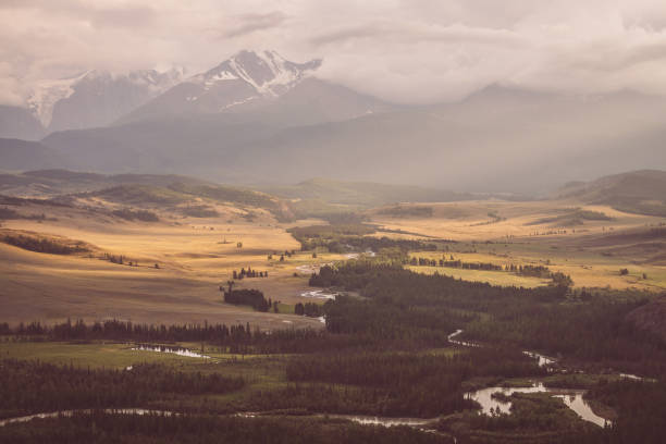 paysage vintage avec vaste plateau avec rivière de montagne et forêt sur fond de crête de montagne enneigée sous un ciel nuageux. vallée de montagne et chaîne de montagnes parmi les nuages bas dans les tons sépia et fanés. - russia river landscape mountain range photos et images de collection