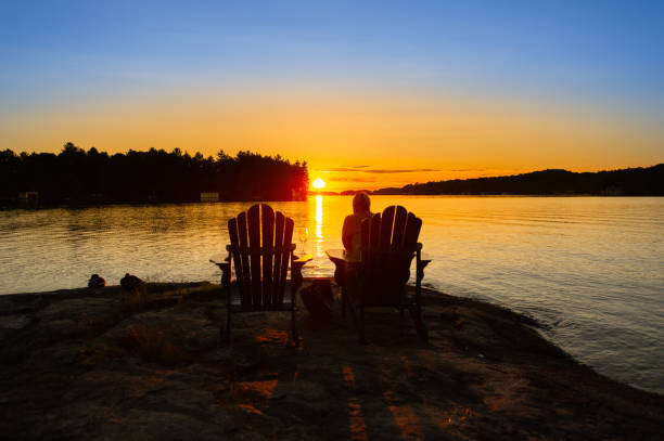 Young woman relaxing in a beautiful sunset setting Young woman relaxing on an Adirondack chair in a beautiful sunset setting. Summer evening on a lake in Muskoka cottage country, Canada. cottage stock pictures, royalty-free photos & images