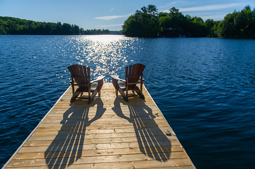 Cottage life - Sunrise on two empty Adirondack chairs sitting on a dock on a lake in Muskoka, Ontario Canada. The sun light is reflecting on the water.