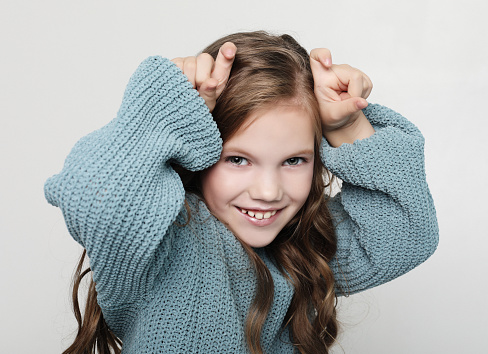 cute girl plays with friends. Portrait of positive happy child with curly hair, holding index fingers above head, mimicking horns and smiling broadly, standing over white background close up