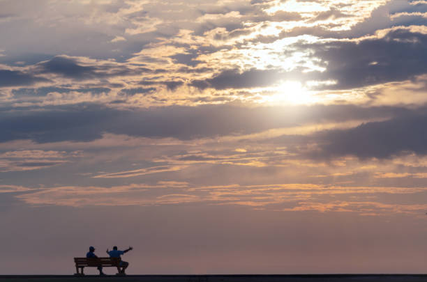 два человека сидят на уличной скамейке и наблюдают за драматическим закатом - romance lake chair sunset стоковые фото и изображения