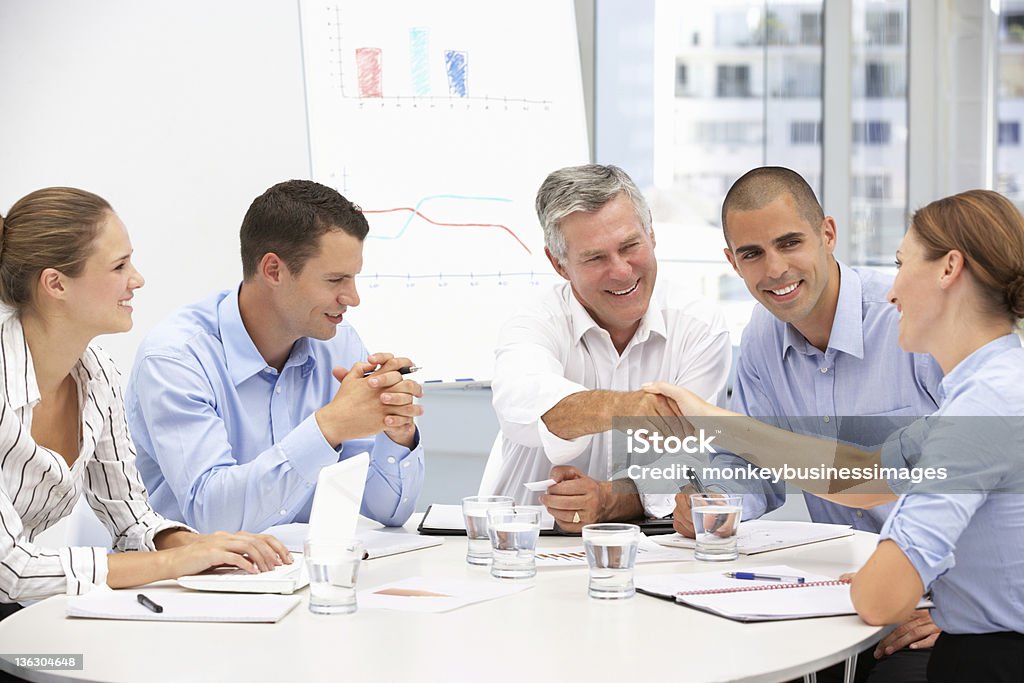 People sitting around a table with papers Business people in a group meeting shaking hands Old Stock Photo