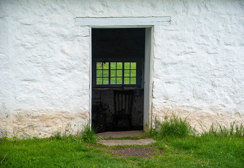 An eighteen-pane colonial window is visible with green grass beyond through open door of a whitewashed stone colonial spring house. Green grass around the stone threshold and facade with white stone texture. Natural light, no people with copy space.