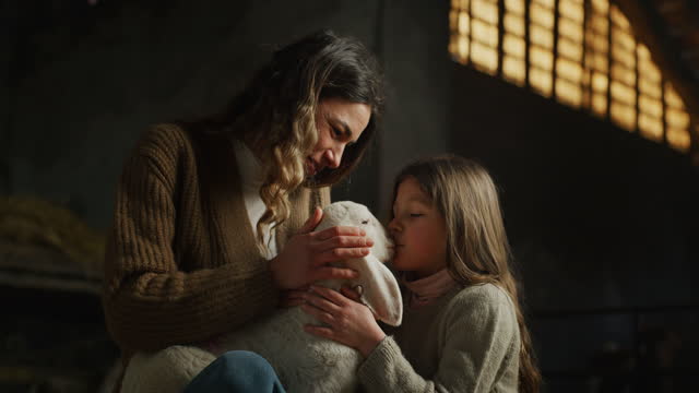 Cinematic shot of happy mother and little daughter caressing with love and care ecologically grown newborn lamb used for biological genuine wool industry in hay barn of countryside agricultural farm.