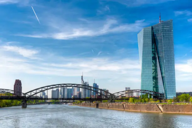 The building of the European Central Bank (ECB) in Frankfurt am Main with the city skyline in the background, a steel arch bridge, quay walls and the river Main in the foreground