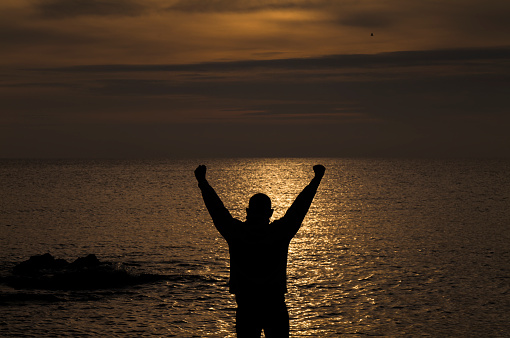 Silhouette of adult man raising arms looking at sea during sunset. Almeria, Spain