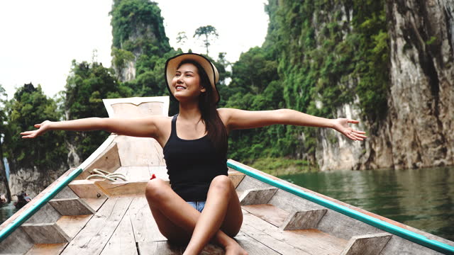 Cheerful Asian young woman wear hat sitting on the wood long tail boat in tropical with raised hands relax over nature background. Travel adventure, Freedom, experience, sightseeing concept.