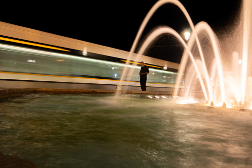 A man walking his dog near a Fountain in Downtown District of Valencia, Spain, with a metro train moving past