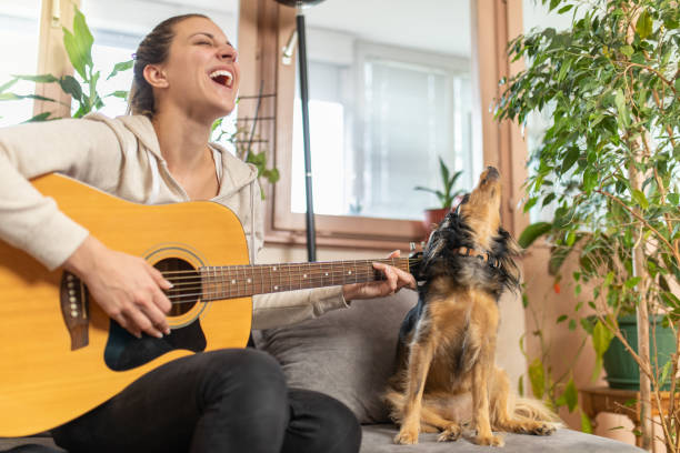 молодая женщина играет на гитаре, пока ее собака поет - 20s acoustic guitar adult attractive female стоковые фото и изображения