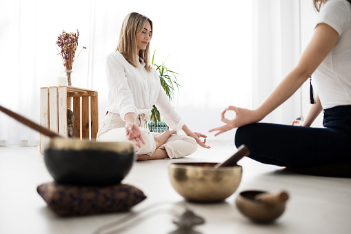 A yoga teacher and her student meditate sitting across from each other. There are various objects such as Tibetan bowls, a Buddha figure and dried flowers.