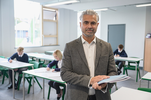 Grey-haired mixed race man with facial hair standing at front of secondary school classroom holding composition booklets and looking at camera with contented expression.