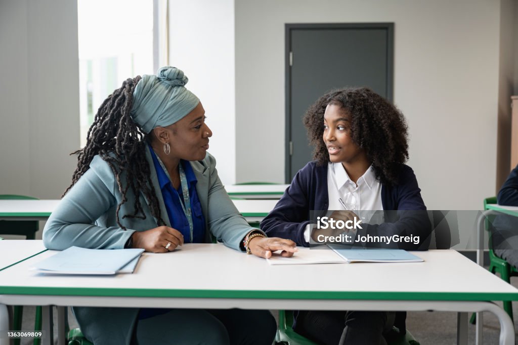 Black educator working with multiracial student in classroom Front view of supportive female teacher in early 50s sitting at desk with teenage schoolgirl while discussing her writing assignment. Teacher Stock Photo