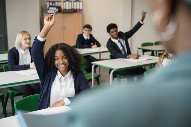 Smiling teenagers participating in classroom discussion Personal perspective over the shoulder of female educator as she faces multiracial group of secondary students in uniforms with hands raised. hand raised classroom student high school student stock pictures, royalty-free photos & images