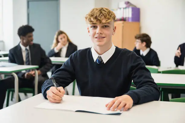 Candid portrait of blond boy in shirt, necktie, and sweater sitting at desk in secondary classroom and smiling at camera while classmates talk in background.