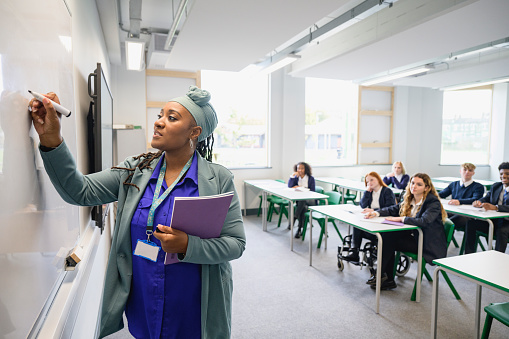 Side view of female educator teaching class as teenage students in uniforms sit at desks in background watching and listening.