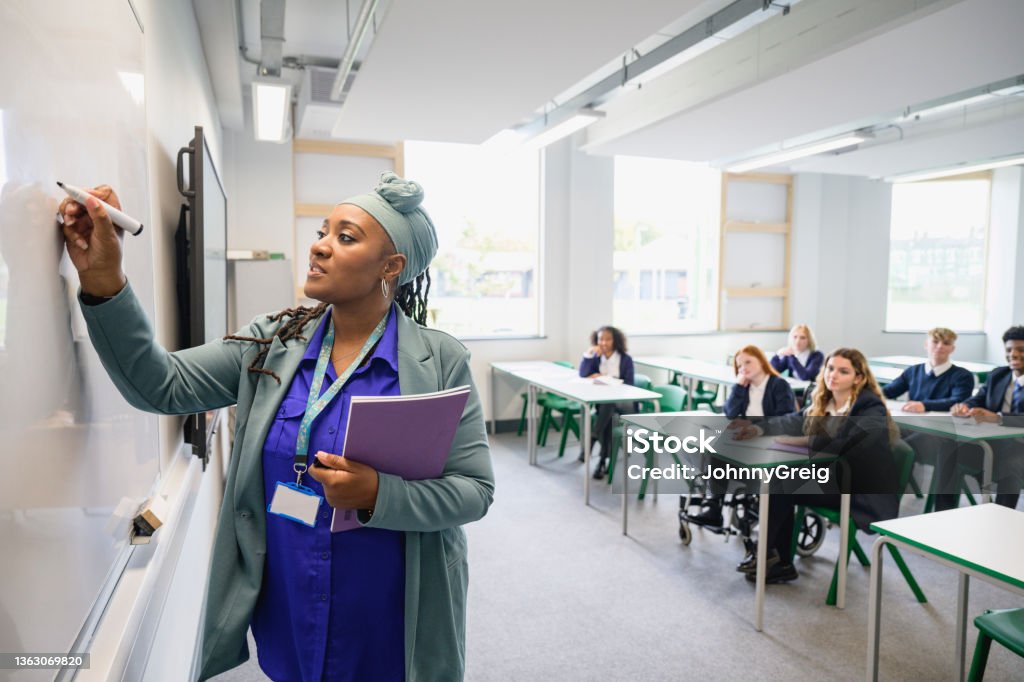 Mature Black instructor writing on whiteboard Side view of female educator teaching class as teenage students in uniforms sit at desks in background watching and listening. Teacher Stock Photo