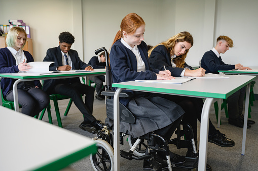 Three-quarter front view of teenage schoolgirl in wheelchair and classmates writing their exams in composition booklets.