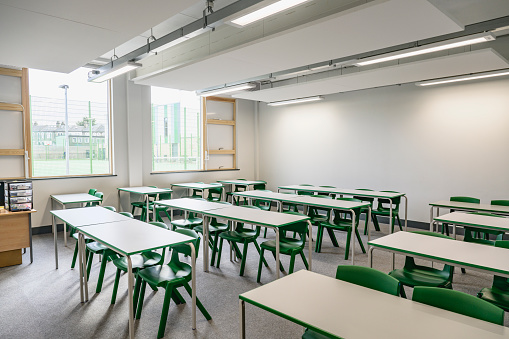 Wide angle view of unoccupied educational environment with desks accommodating two students each, chairs, windows, and overhead LED lighting.