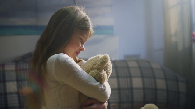 Cinematic shot of cute little girl is telling a magic fairy tale to her teddy bear and hugging it while sitting on a cosy carpet in a nursery. Concept of love, childhood, friendship and sweet dreams.