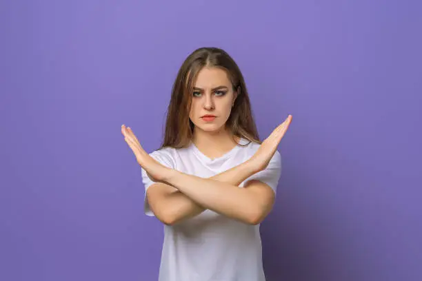 Serious girl showing stop cross gesture, disapprove something, prohibit action, make taboo no sign, standing over purple background. Young emotional woman in purple hoodie