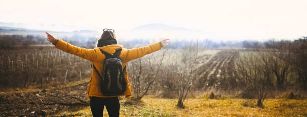 Backpacker woman tourist traveling alone and posing at dawn with fog on beautiful nature with mountains in autumn. Banner copy space. Blonde lady in yellow down jacket back view with arms outstretched stock photo