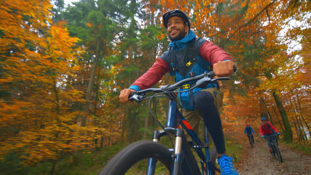 Wide tracking shot of a young man riding his mountain bike on a gravel road through the fall forest and his friends are riding their bikes behind him. Shot in Slovenia.
