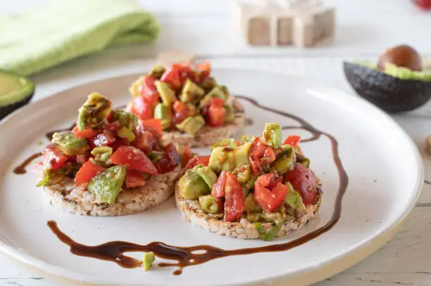 Healthy gluten free and vegetarian meal with a avocado salad with chopped tomatoes, balsamic dressing. Served on brown rice cracker on white table background.