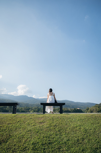 Back view of woman in white dress sitting on seat at hill and blue sky on sunny day in public park