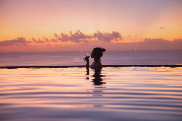 Asian Woman Enjoying Wine in an Infinity Pool at Sunset Sideview shot of an unrecognizable rich Asian woman drinking a glass of wine while enjoying sunset inside an infinity pool of a luxury resort. infinity pool stock pictures, royalty-free photos & images