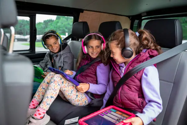 A side-view shot of a group of young triplets sitting in the back of their parent's car, they are wearing headphones and using digital tablets to play games, they are smiling and having fun.