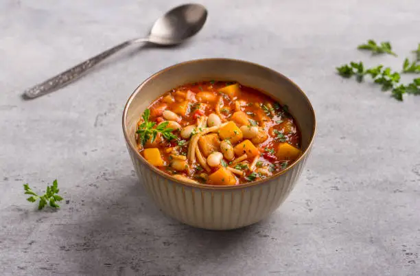 Vegan seasonal italian minestrone soup with pumpkin, beans, tomatoes, bell peppers, pasta and herbs on gray textured background. Delicious homemade food