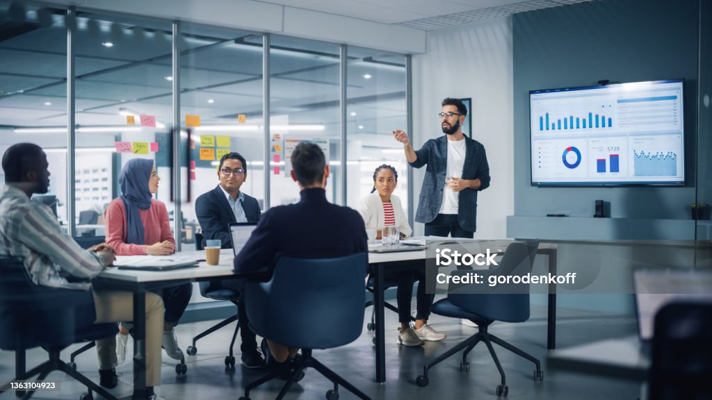 Diverse Modern Office: Businessman Leads Business Meeting with Managers, Talks, uses Presentation TV with Statistics, Infographics. Digital Entrepreneurs Work on e-Commerce Project. Business Stock Photo