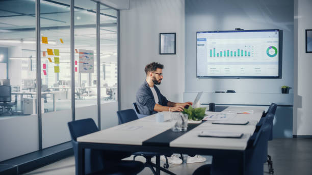 istock Office Conference Meeting Room: Handsome Hispanic Specialist Working on Laptop Computer with Big Wall TV with Big Data Analysis, Charts and Infographics. Businessperson Working in Growing Startup. 1363105059