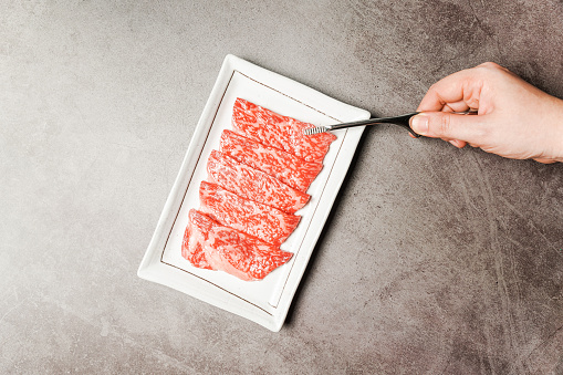 Premium Rare Slices sirloin Wagyu A5 beef with high-marbling texture on a ceramic plate, pick up by bbq tongs with hand. Served for Yakiniku, Sukiyaki and Shabu. Image with copy space.