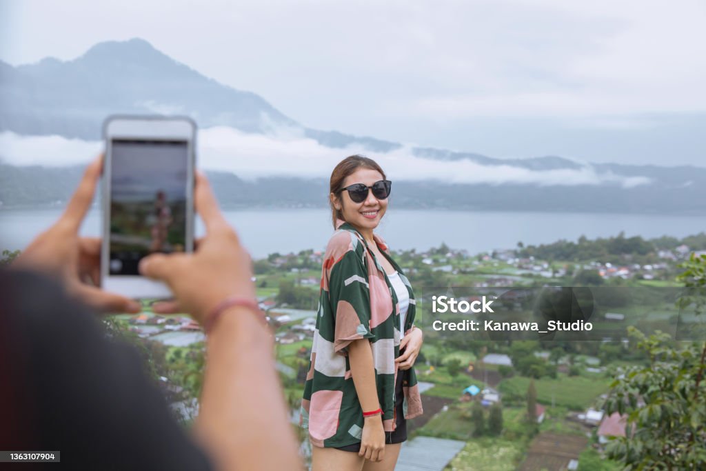 Insta Boyfriend Photographing Girlfriend with Mountain View Shot of an unrecognizable insta boyfriend taking a picture of his girlfriend using smartphone with Batur mountain view behind her during their weekend getaway in Bali Kintamani Stock Photo