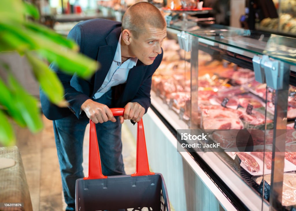Focused man examines the production in the department of the butcher shop in the supermarket Focused man with a grocery basket carefully examines the production in the department of the butcher shop in the 
supermarket 30-34 Years Stock Photo